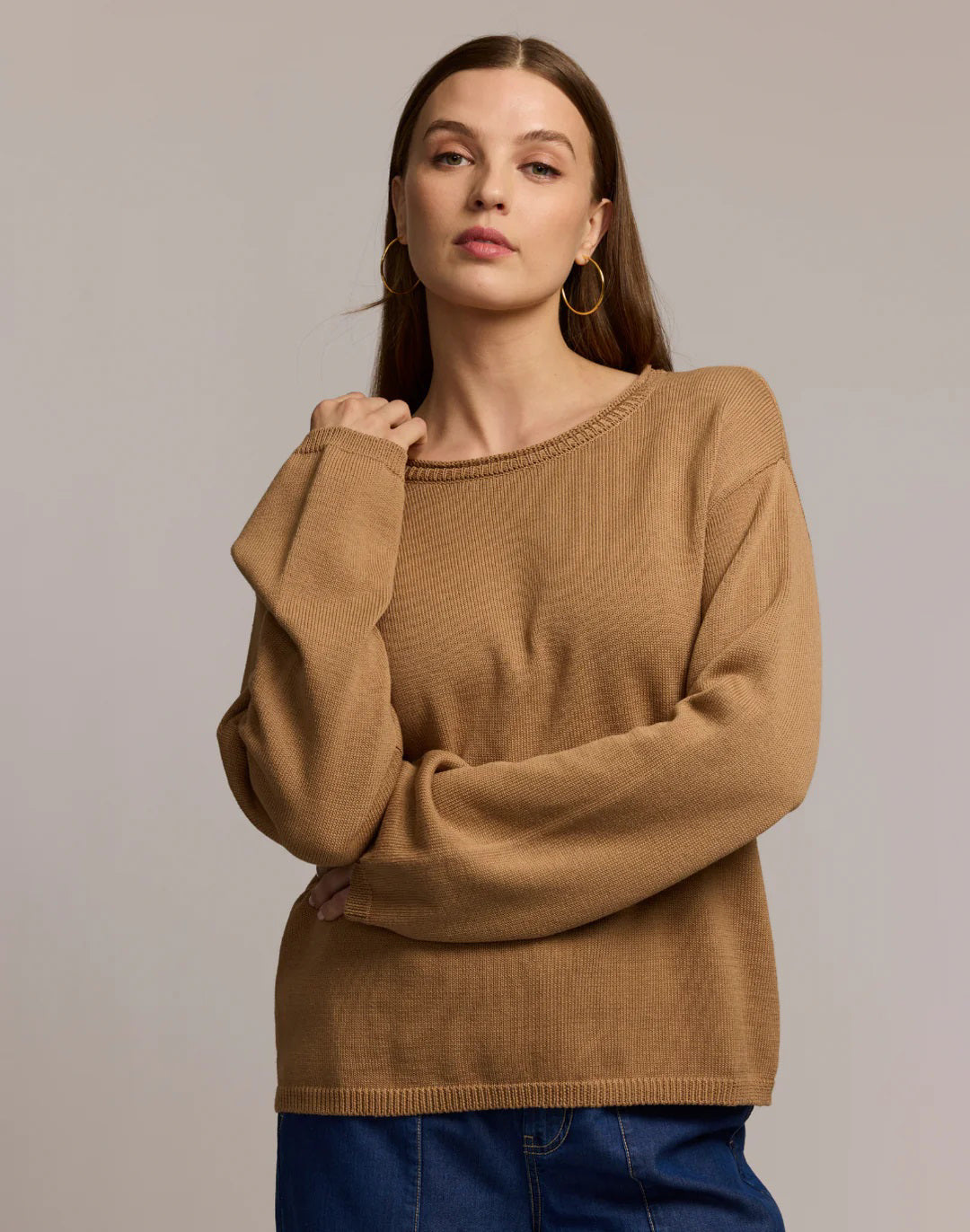 A woman with long brown hair and large hoop earrings models the HINSON WU AUDREY Long Sleeve Cotton Silk Sweater in tan with blue jeans, standing against a plain beige background, one hand raised near her shoulder.