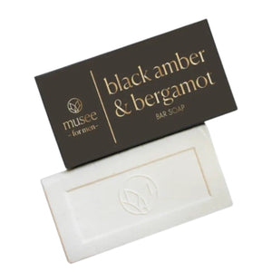 A rectangular MUSEE - BLACK AMBER & BERGAMOT BAR SOAP in a brown box labeled with the product name rests on a wooden surface beside a green potted succulent. Crafted by MUSEE, this moisturizing bar soap brings elegance and a fresh aroma to any space.