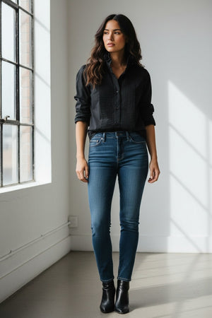 A blonde older woman wears the KMJ - SOUTHERN LOVER TUX blouse by KMJ and blue demi-boyfriend jeans with a turquoise necklace, looking down and tucking her hand in her waistband, set against a plain white background.