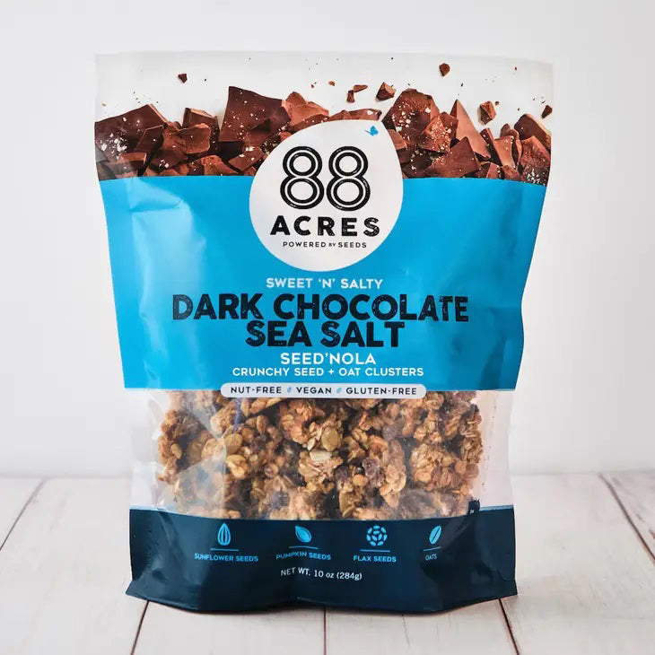 A bag of 88 ACRES - DARK CHOCOLATE SEA SALT SEED'NOLA sits on a light surface. Its blue and white packaging highlights vegan, nut-free seeds, gluten-free oats, and dark chocolate with granola imagery.