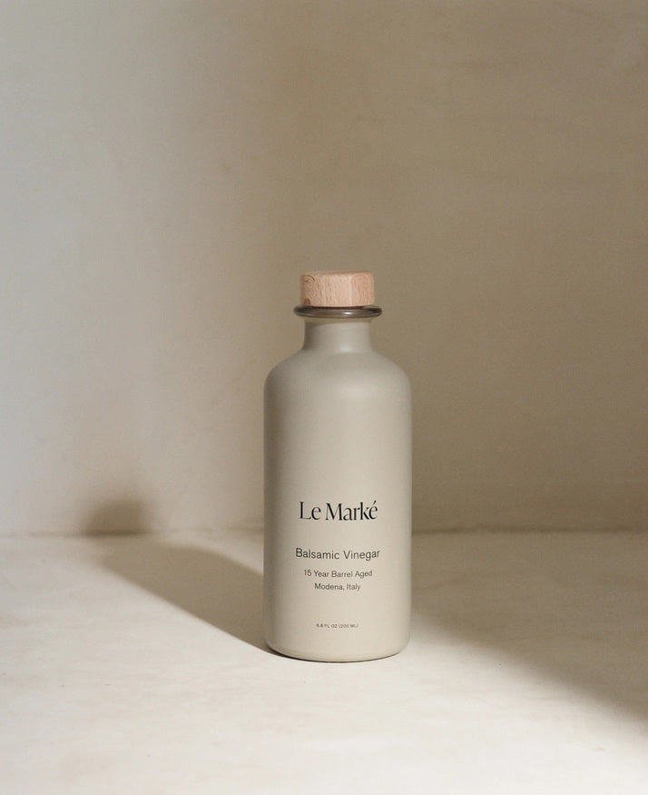 A minimalist bottle of LE MARKE - BALSAMIC VINEGAR, crafted from Trebbiano grapes, stands against a neutral background. The beige bottle, adorned with a wooden cork stopper and a label that reads &quot;LE MARKE Balsamic Vinegar 18 Year Barrel Aged Modena, Italy,&quot; exudes elegance and tradition.