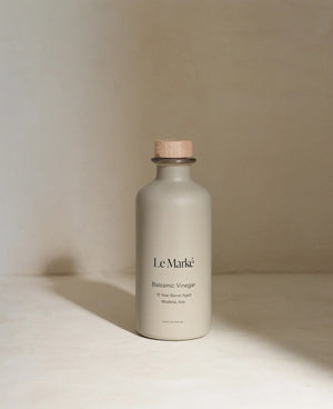 A minimalist bottle of LE MARKE - BALSAMIC VINEGAR, crafted from Trebbiano grapes, stands against a neutral background. The beige bottle, adorned with a wooden cork stopper and a label that reads "LE MARKE Balsamic Vinegar 18 Year Barrel Aged Modena, Italy," exudes elegance and tradition.