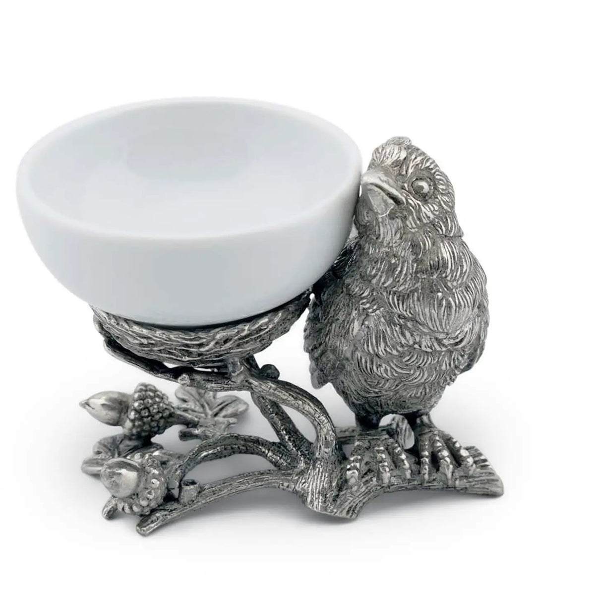 A small white bowl of butter sits on Vagabond House&#39;s Songbird Salt Cellar, showcasing intricate pewter designs with delicate feathers. The scene is set on a dark surface with blurred greenery in the background.