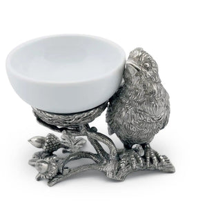 A small white bowl of butter sits on Vagabond House's Songbird Salt Cellar, showcasing intricate pewter designs with delicate feathers. The scene is set on a dark surface with blurred greenery in the background.