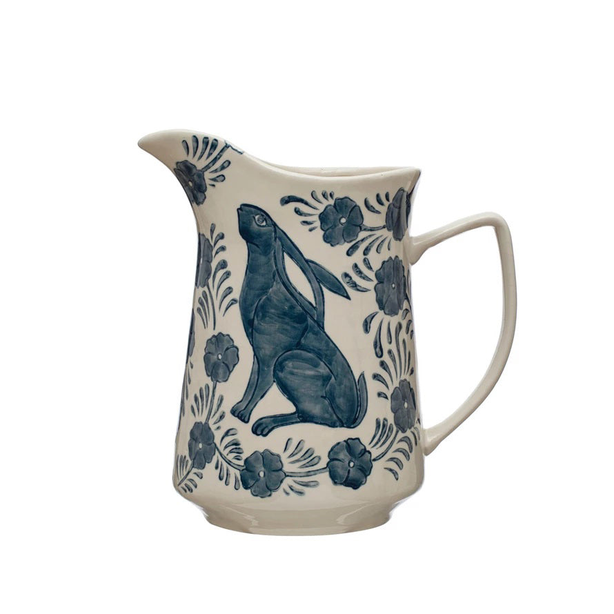 The HAND PAINTED STONEWARE PITCHER WITH RABBIT &amp; FLOWERS from CREATIVE COOP features a charming blue design of a rabbit seated on its hind legs, surrounded by floral and leafy patterns. This 3-quart white and blue ceramic pitcher, complete with spout and handle, beautifully showcases the intricate Rabbit &amp; Flowers decorations.