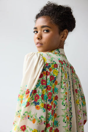 A woman with curly hair looks over her shoulder, wearing the HOPE FOR FLOWERS Smocked Tie Neck Blouse in Custard Floral—a sustainable, cream-colored top with vibrant florals from HOPE FOR FLOWERS, set against a plain, softly lit background.