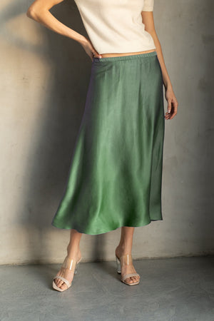 An older woman with long gray hair stands smiling in a dark red ROSE & CROWN cardigan, the ROSE AND CROWN - SATIN MIDI SKIRT, and black knee-high boots against a plain white background.