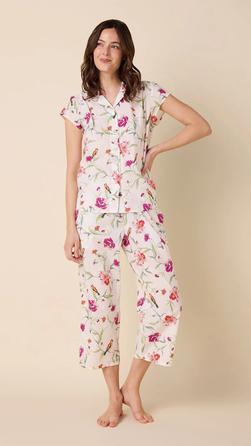 A person stands barefoot, wearing the CAT&#39;S PAJAMAS - ROSA AND BIRD LUXE PIMA CAPRI PAJAMA SET from CAT&#39;S PAJAMAS, featuring pink, purple, and green florals on a cream 100% cotton base with short sleeves and cropped pants. The backdrop is light beige.