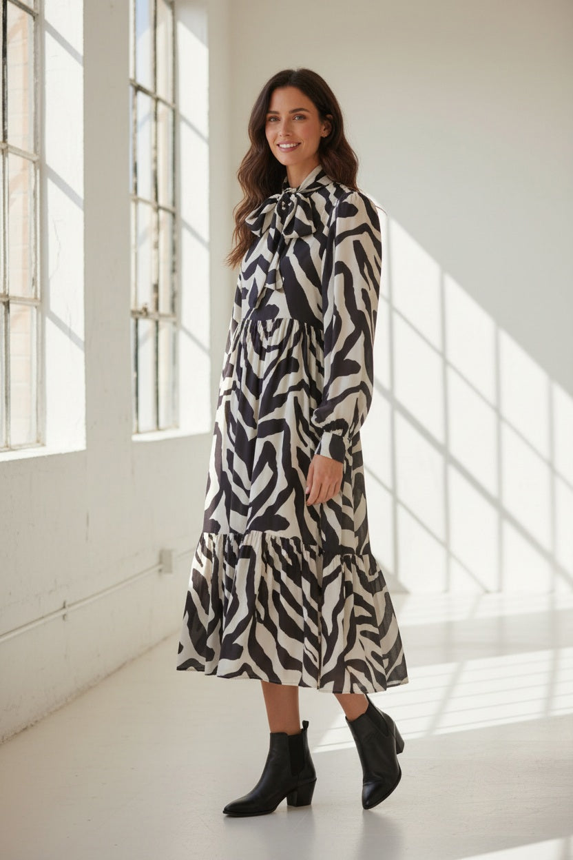 A person is seen from behind wearing the MEIMEIJ RUFFLE PRINT DRESS—a long-sleeve, black-and-white zebra print design crafted from 100% viscose by MEIMEIJ—set against a light, textured background.