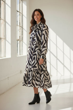 A person is seen from behind wearing the MEIMEIJ RUFFLE PRINT DRESS—a long-sleeve, black-and-white zebra print design crafted from 100% viscose by MEIMEIJ—set against a light, textured background.