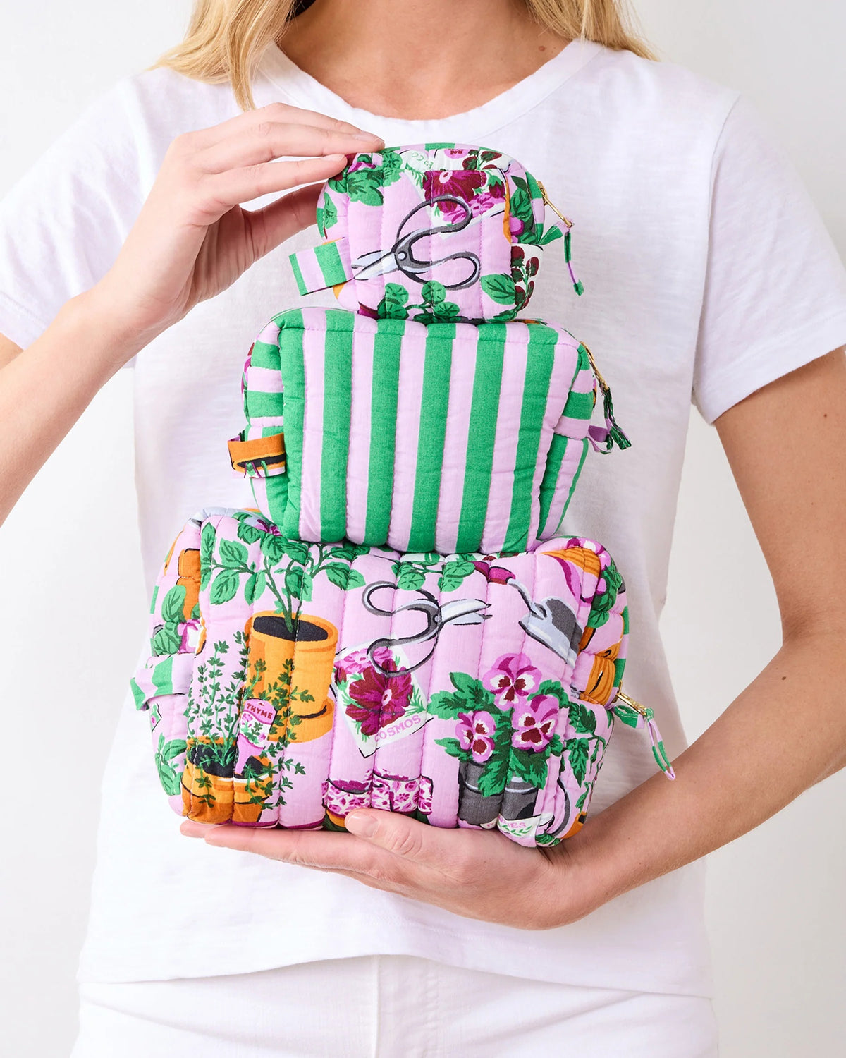 A person in a white shirt holds the PRINTFRESH - GREEN THUMB THREE POUCH SET by PRINTFRESH, featuring three quilted travel pouches with floral and striped patterns, stacked from largest to smallest, against a plain background.