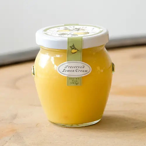 A jar of zesty BELLA CUCINA - LEMON CREAM with a white lid, is beautifully adorned with a green ribbon featuring a small lemon graphic. The label on the front, in elegant lettering, reads "BELLA CUCINA - LEMON CREAM," and the jar is set against a simple white background.