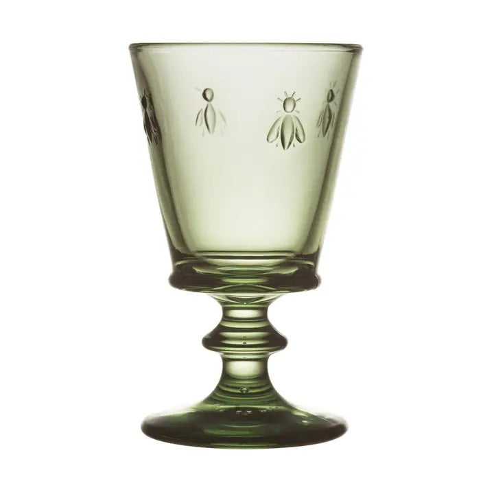A table set for a meal features LA ROCHERE’s BEE WINE GLASS in Olive, along with clear glasses, plates, cutlery, and decorative greenery atop a white tablecloth.
