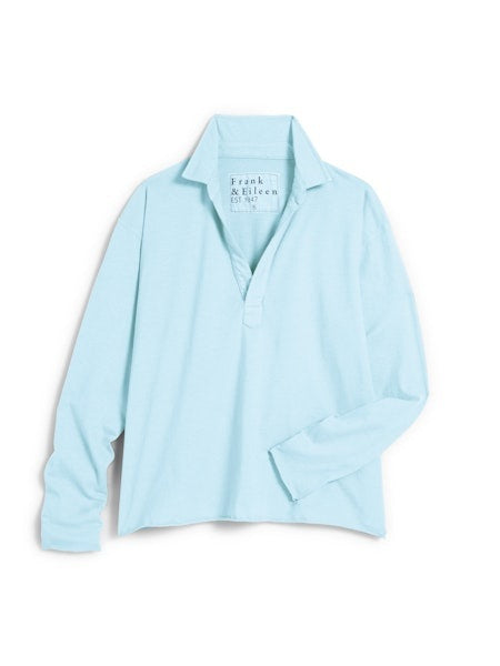 A woman with wavy brown hair smiles in a bright, minimalist room, wearing the FRANK &amp; EILEEN Patrick Popover Henley in Vintage Jersey Dive, blue jeans, and white sneakers, with one hand casually in her pocket.