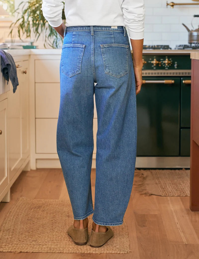 A person stands in a kitchen with their back to the camera, wearing FRANK &amp; EILEEN’s Brennan Everyday Barrel Jean in 1978 Vintage Wash, a white long-sleeve shirt, and brown shoes. The kitchen has light cabinets, a dark oven, and wood floors.