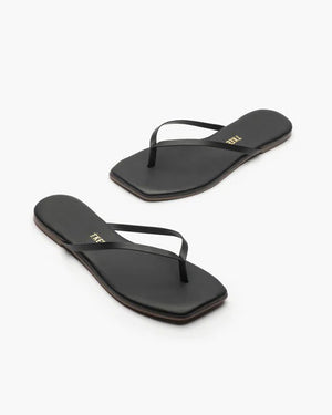 The TKEES INC SQUARE TOE LILY BLACK sandals feature a black leather upper with thin straps and square toes, shown from above on a white background. "TKEES" is printed in gold on the insoles, emphasizing their modern design.