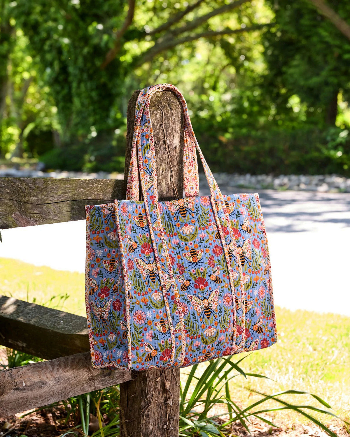 The PRINTFRESH - Bee Tapestry Traveler Tote in Delphinium Blue by PRINTFRESH, featuring vibrant floral, butterfly, and bee designs on organic cotton, hangs on a rustic wooden fence outdoors with lush green foliage in the background.
