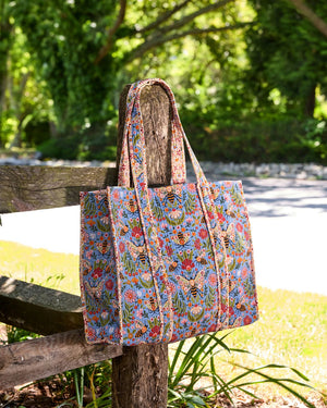 The PRINTFRESH - Bee Tapestry Traveler Tote in Delphinium Blue by PRINTFRESH, featuring vibrant floral, butterfly, and bee designs on organic cotton, hangs on a rustic wooden fence outdoors with lush green foliage in the background.