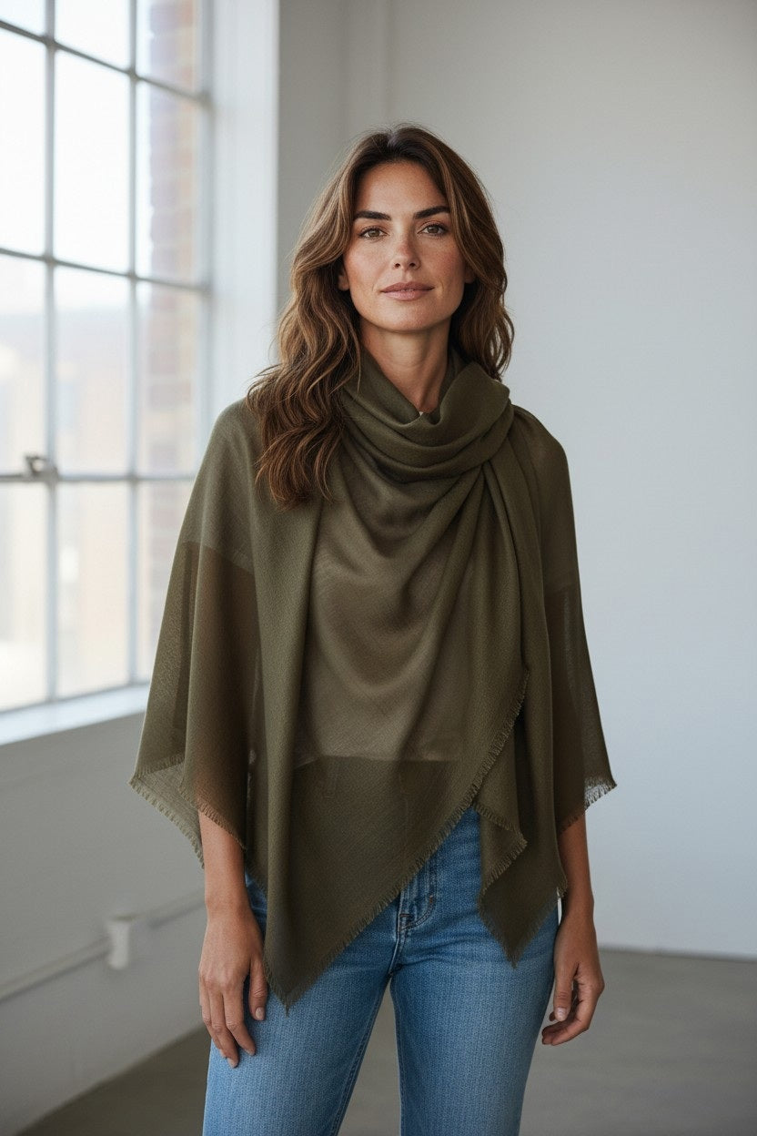 A woman with wavy brown hair, wearing the FRANCO FERRARI Cashmere Scarf 135x135 in green and blue jeans, stands indoors near large windows with natural light.