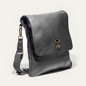 The WILL LEATHER GOODS OTTO Crossbody Bag in black full-grain leather features a front flap, gold-tone hardware, adjustable shoulder strap, and a small stitched patch on the lower right corner. Displayed against a plain white background.