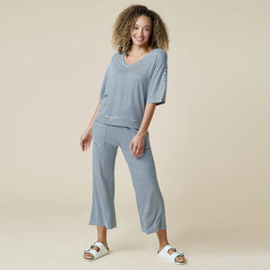 A woman wearing a light blue and white striped SOFTIES - DREAM RELAXED V-NECK WITH CAPRI LOUNGE SET with a V-neck, capri-length pants, and white sneakers.