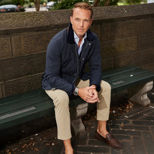 A man, face unseen, stands by a light wall with hands in pockets, wearing a light blue shirt, COLLARS AND CO - APEX TECH PANTS IN LIGHT BEIGE featuring four-way stretch, a brown belt, and brown loafers.