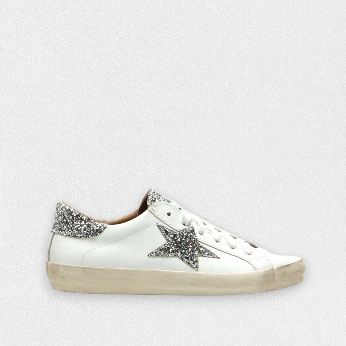 The MICHELE LOPRIORE - GEMMA SNEAKER WITH SILVER STAR features a white low-top design with a glittery silver star on the side and a matching heel tab. Made from soft leather and sporting an extra light rubber sole, it has a stylishly worn look with a beige sole and white laces against a plain, textured light grey background.