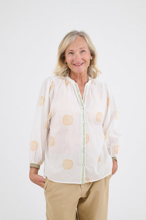 A smiling older woman with blonde hair stands against a white background, hands in her pockets, wearing the STORIATIPIC - LUCIE SOLEIL BLOUSE by STORIATIPIC, featuring orange patterns for a look of casual elegance.