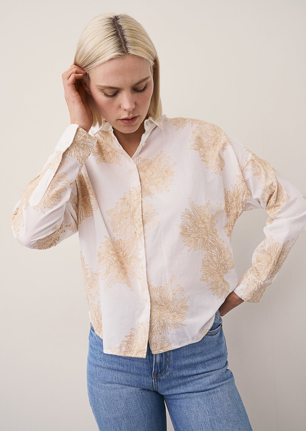 A woman with straight blonde hair and a neutral expression stands against a plain light wall, wearing the 0039 ITALY LUCY BLOUSE WITH EMBROIDERED FLOWERS, light blue wide-leg jeans, and beige flat shoes.