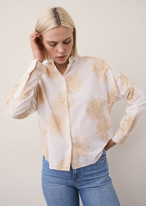 A woman with straight blonde hair and a neutral expression stands against a plain light wall, wearing the 0039 ITALY LUCY BLOUSE WITH EMBROIDERED FLOWERS, light blue wide-leg jeans, and beige flat shoes.