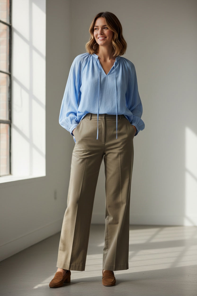 A person wearing the ELEH - BLOUSE WITH EMBRIODERY by ELEH, a loose, light blue peasant-style blouse with long sleeves and tie details, paired with white shorts, stands against a plain light background.