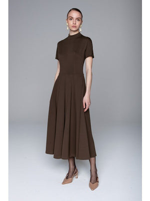 A woman faces away, wearing the LAVI COUTURE - MIDI DEIOPEA DRESS by LAVI in brown. The minimalist design features short sleeves and a fitted waist, styled with a neat bun and heeled sandals against a plain gray background.