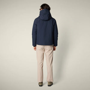 A man in sunglasses, beige wide-leg pants, and olive green shoes wears the SAVE THE DUCK - FLYNN HOODED JACKET by SAVE THE DUCK. He stands against a light background facing forward, with one hand by his side and one near his zipper.