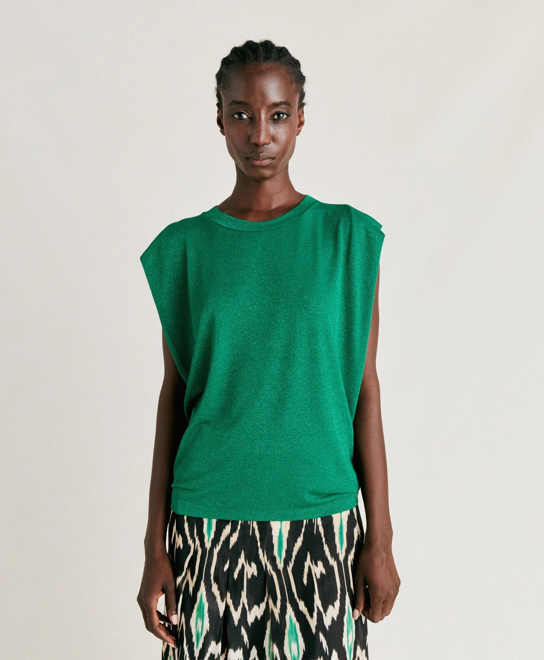 A person with braided hair is wearing a MOMONI ENNA LUREX JERSEY TOP and a skirt featuring a black, white, and green patterned design reminiscent of an 80s interpretation. The background is plain and neutral.