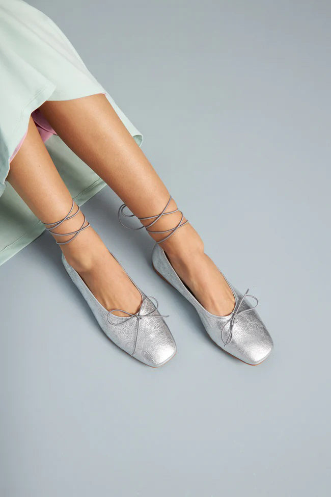 The Augusta - Sienna Ballet Flat by AUGUSTA is a single silver-colored shoe featuring a subtle metallic sheen and a simple bow on the front. It has a low brown sole and a pull tab at the back. Handcrafted in Spain from leather, this ballet flat is shown from the side against a light gray background.