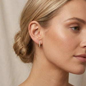 A woman with blonde hair styled in a low bun wears the ELLA STEIN - HALF HEARTED ADVENTURE NEVER HOOP EARRINGS from ELLA STEIN, featuring small gold plated hoops adorned with sparkling stones. The photo captures her profile against a neutral background.