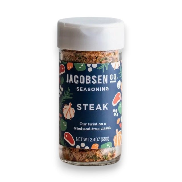 A 2.4 oz (68g) jar of JACOBSEN SALT CO. JACOBSEN SALT - STEAK SEASONING features a blue label decorated with illustrations of garlic, herbs, and meat. Expertly crafted with kosher salt, this seasoning blend is perfect for enhancing your favorite steak cuts.