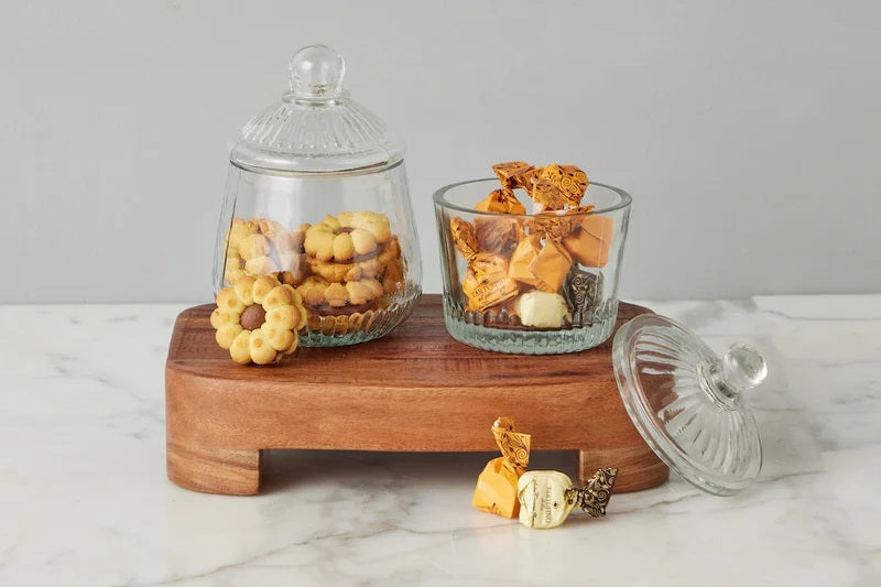 ETUHOME Bistro Glass Biscuit Jars with lids sit on a wooden tray, filled with snacks and orange-wrapped candies. The classic jar design by ETUHOME stands out against the softly blurred background.