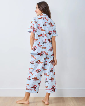 A woman with long, wavy brown hair stands barefoot against a white wall, wearing the PRINTFRESH Dachshund Date Short Sleeve Top & Cropped Pants Pajama Set in Blue. The organic cotton set from PRINTFRESH features short sleeves and cropped pants adorned with a charming dachshund print. She smiles slightly.