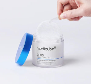 A hand holds a round, textured exfoliating pad above an open jar of KOSMIC KBEAUTY SKINCARE MEDICUBE - ZERO PORE PAD 2.0, 100ML, with its blue lid set aside on a plain white background.