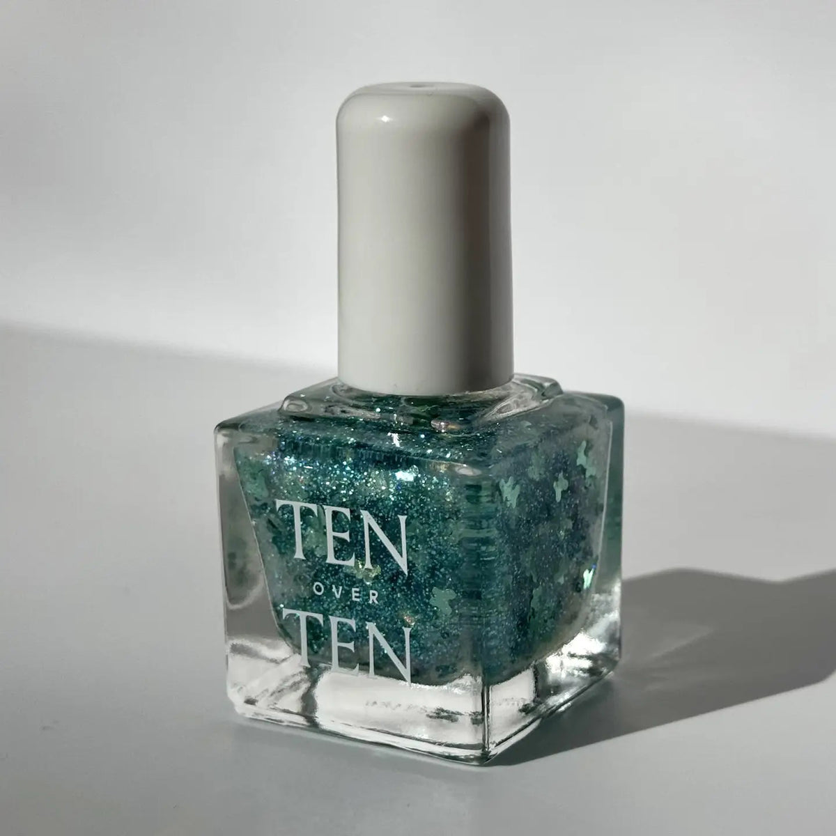 A glass bottle of TENOVERTEN - NAIL POLISH from the brand TEN OVER TEN, featuring a white cap and a dusty pink nail color. The front label displays "TEN OVER TEN" in white uppercase letters. This vegan, eight-free formula ensures both beauty and peace of mind. The background is plain and light-colored.