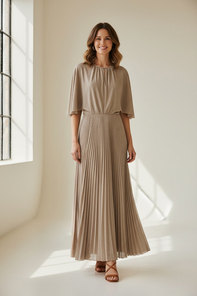 A woman poses against a plain background wearing the MOMONI - PARADISU DRESS by MOMONI, featuring light beige lurex jersey, kimono sleeves, and a pleated skirt. She styles it with braided hair, earrings, and open-toed shoes.