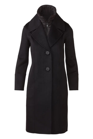 A woman stands indoors by a large window, smiling at the camera, wearing blue jeans and the KINROSS CASHMERE Quilted Insert Notch Collar Coat in black layered over a quilted black jacket.