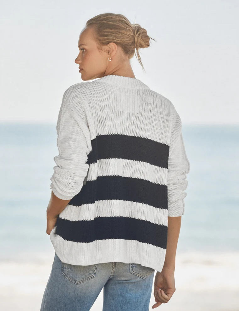 A woman stands on a beach wearing the FRANK & EILEEN Montecito Crew Neck Cardigan in Pure Italian Cotton, featuring navy and white stripes in a relaxed fit with large buttons, paired with light blue jeans. The ocean and sky are softly blurred in the background.