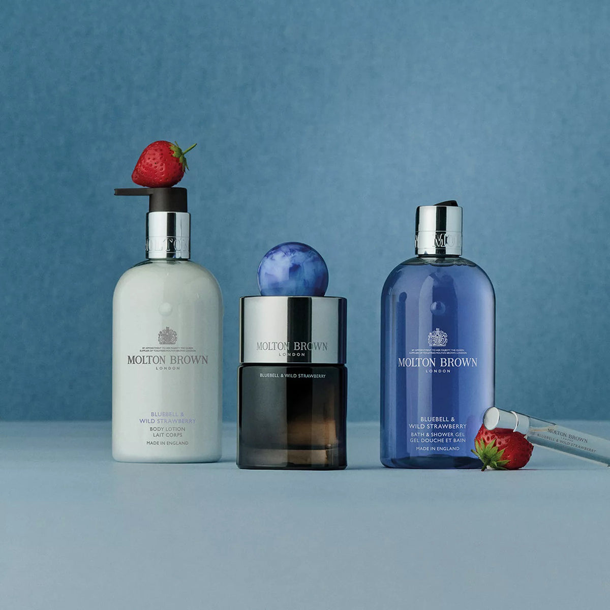 Three MOLTON BROWN Bluebell &amp; Wild Strawberry Body Wash bottles are displayed on a light blue surface with a blue background, surrounded by strawberries and an applicator, evoking the floral-fruity fragrance to entice your senses.