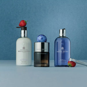 Three Molton Brown Bluebell & Wild Strawberry Eau de Parfum bottles sit on a blue surface with fresh strawberries, emphasizing the collection’s wild strawberry and floral-fruity scent. A silver tube is placed on the right side.
