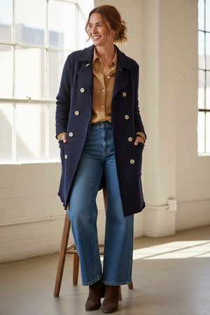 A woman stands indoors by a window, smiling with her hands in the pockets of a FRANK & EILEEN Belfast Sweatshirt Trench Coat in Classic Navy from the California Heritage Collection. She wears a tan shirt, wide-leg blue jeans, and brown boots. A wooden stool is positioned behind her.