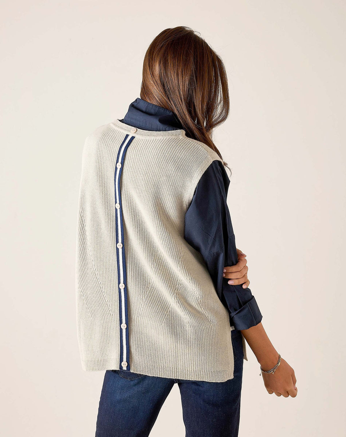 A woman with long brown hair stands sideways against a light background, looking at the camera, wearing the MERSEA - CAMDEN VEST IN DESERT PALM WITH STRIPED TAB by MERSEA over a dark collared shirt and blue jeans.