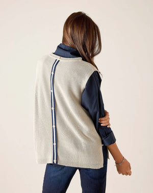 A woman with long brown hair stands sideways against a light background, looking at the camera, wearing the MERSEA - CAMDEN VEST IN DESERT PALM WITH STRIPED TAB by MERSEA over a dark collared shirt and blue jeans.