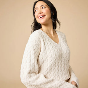 A woman wearing the SOFTIES CABLE KNIT MARSHMALLOW V-NECK WITH FLARED SLEEVE AND PANT SET, a marshmallow-soft, cream-colored cable knit v-neck sweater with wide sleeves and matching lounge pants.
